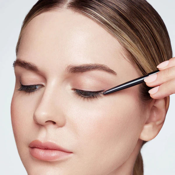 Hold the skin taut to help create a smooth canvas. Starting at the outer corner of the eye, using Defining Liner trace just above the lash line to the inner corner of the eye. Use the smudging tool at the opposite end of the pencil to soften and blend.   